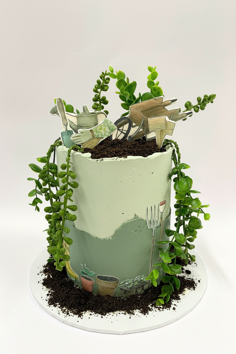 A decorated cake designed to look like a garden with green buttercream and edible plants and gardening tools, including a watering can and rake, with a beige layer at the bottom.