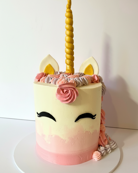 A decorated cake designed to look like a unicorn with a golden horn, pink and white ombre frosting, and ears on top.